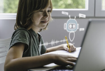 A child at a light wood desk studying a small 3D-printed toy they are deciding how to change, a tablet pushed slightly aside, a sketch notebook nearby, and a clear empty desktop space for the product.